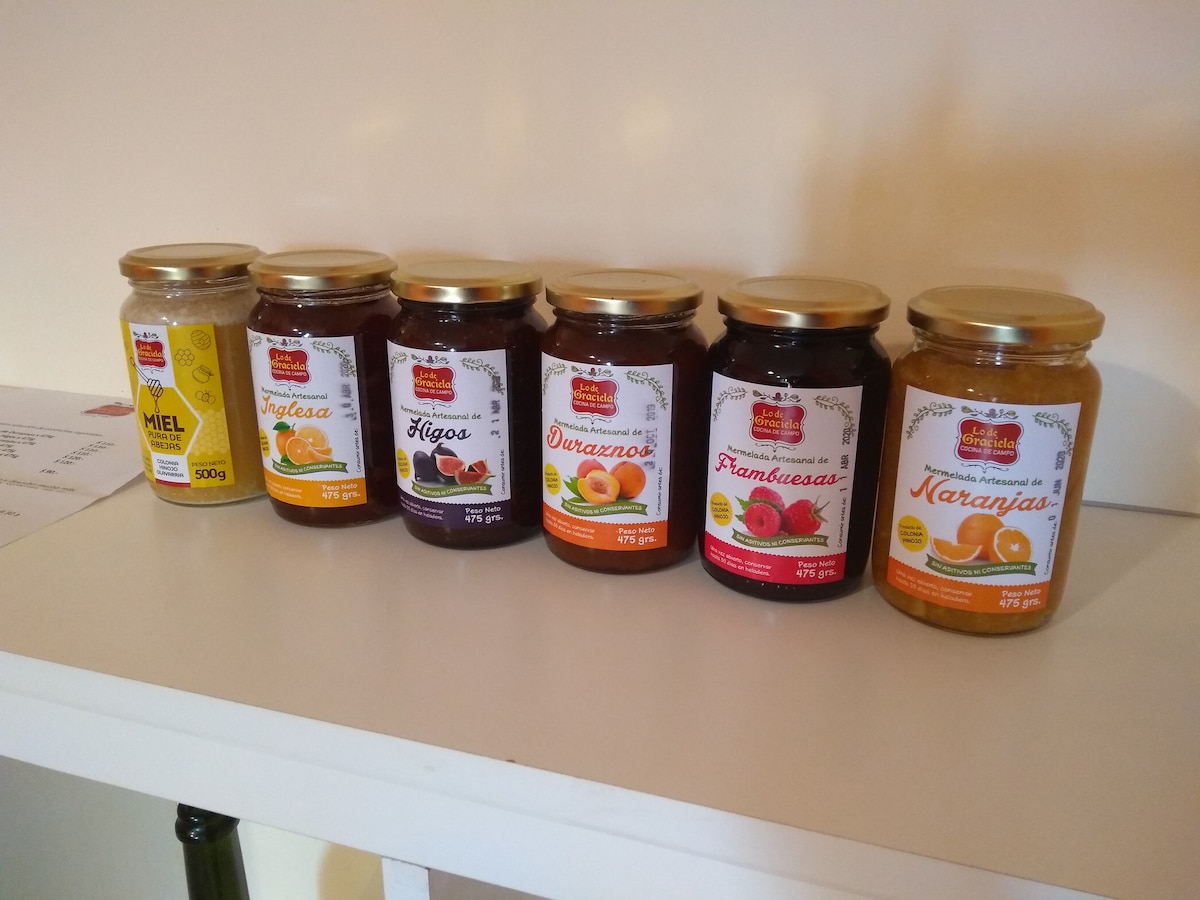 A line of six jars of preserves is displayed on a shelf. Each jar features a golden lid and a label indicating various flavors, including honey, figs, orange, raspberry, and others. The jars are neatly arranged, creating an aesthetically pleasing presentation.