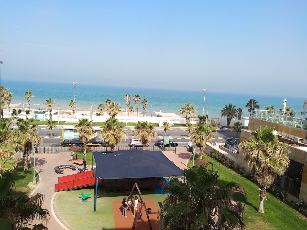 An expansive view of the sea is presented, framed by palm trees swaying gently in the breeze. A colorful playground is visible in the foreground, featuring a shaded area and equipment for children. The sky is clear, creating a bright atmosphere.