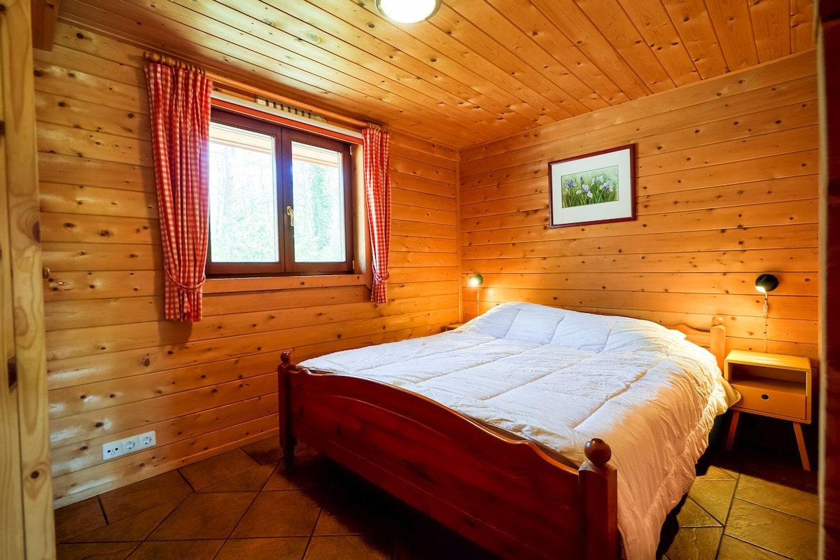 A warm wooden bedroom features a spacious bed with white bedding. A bedside table is located next to the bed, with wall-mounted reading lights enhancing functionality. Natural light enters through a window, framed by red-checkered curtains, while a framed picture adorns the wall.