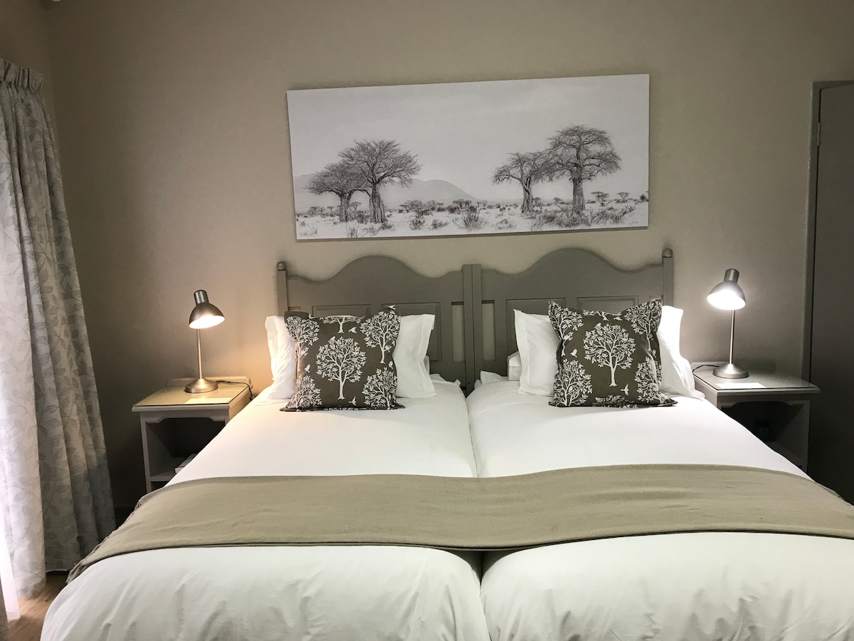 A neutrally decorated bedroom features two twin beds adorned with white linens and decorative pillows. Nightstands with lamps are positioned on either side. A large artwork depicting trees against a light background is displayed above the beds, enhancing the room's tranquil atmosphere.