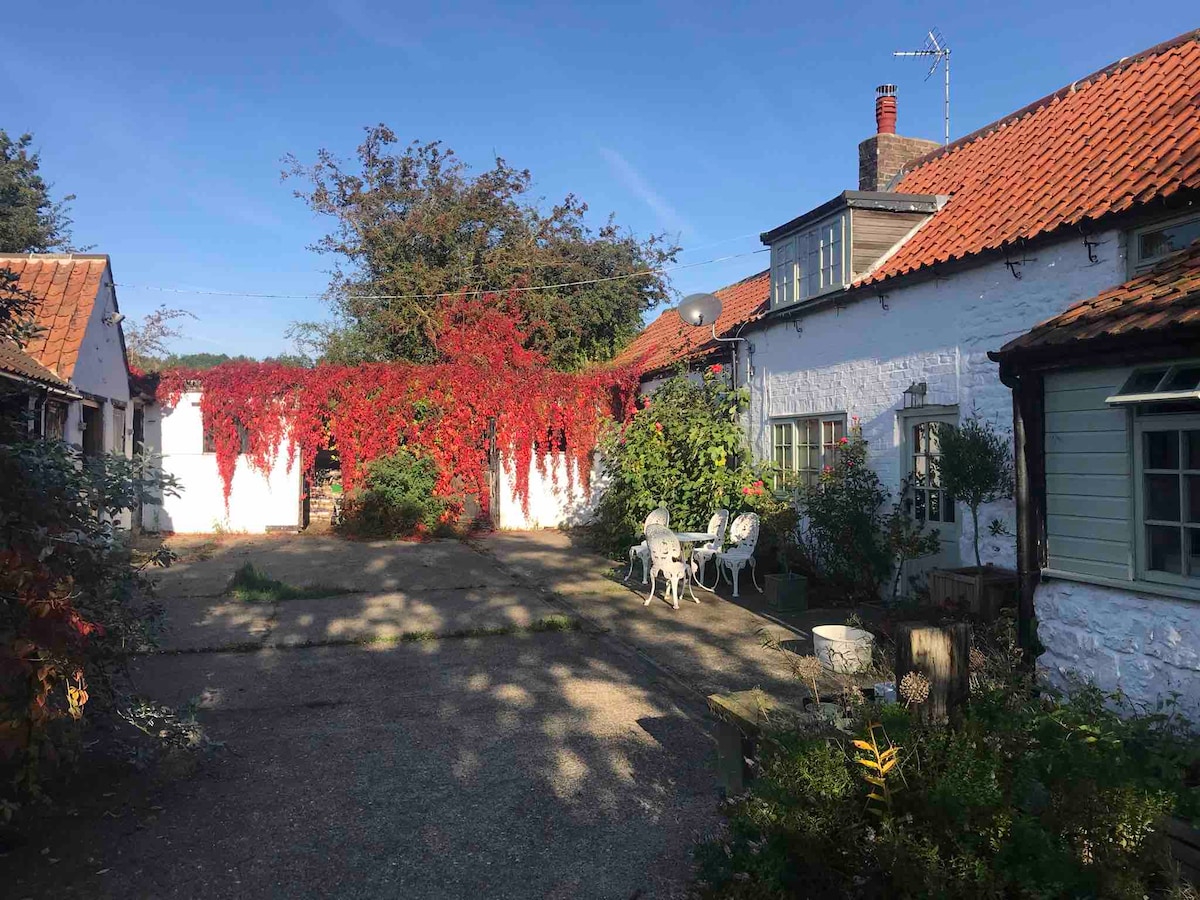 A spacious courtyard is framed by rustic buildings with red brick and whitewashed walls. Vibrant red foliage climbs the exterior, providing a striking contrast to the greenery. A small seating area with white chairs and a table invites relaxation in this tranquil rural setting.