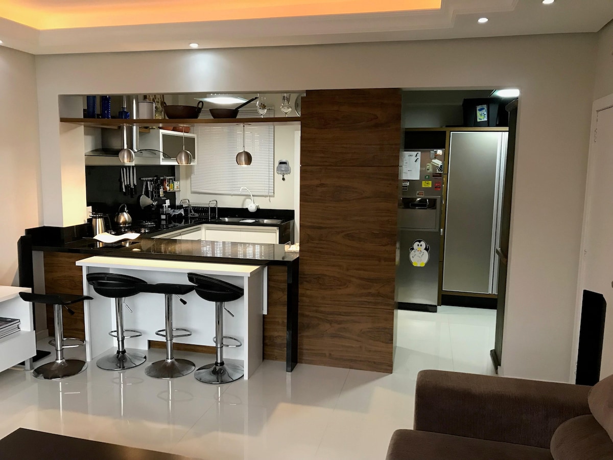 The modern kitchen features sleek cabinetry and an island bar with seating for four. Stainless steel appliances and cookware are organized efficiently, while natural light brightens the space through a window. A refrigerator is positioned adjacent, creating a cohesive cooking area.