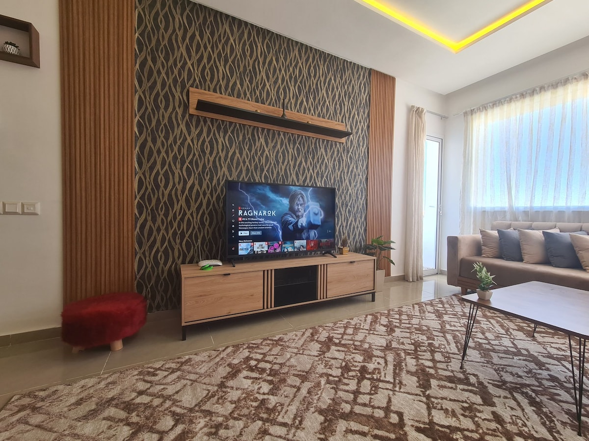 A modern living area features a wooden entertainment unit with a flat-screen TV. A plush sofa with neutral tones is arranged around a low coffee table. Natural light enters through the large windows, which are adorned with sheer curtains. Decorative plants add a touch of greenery.