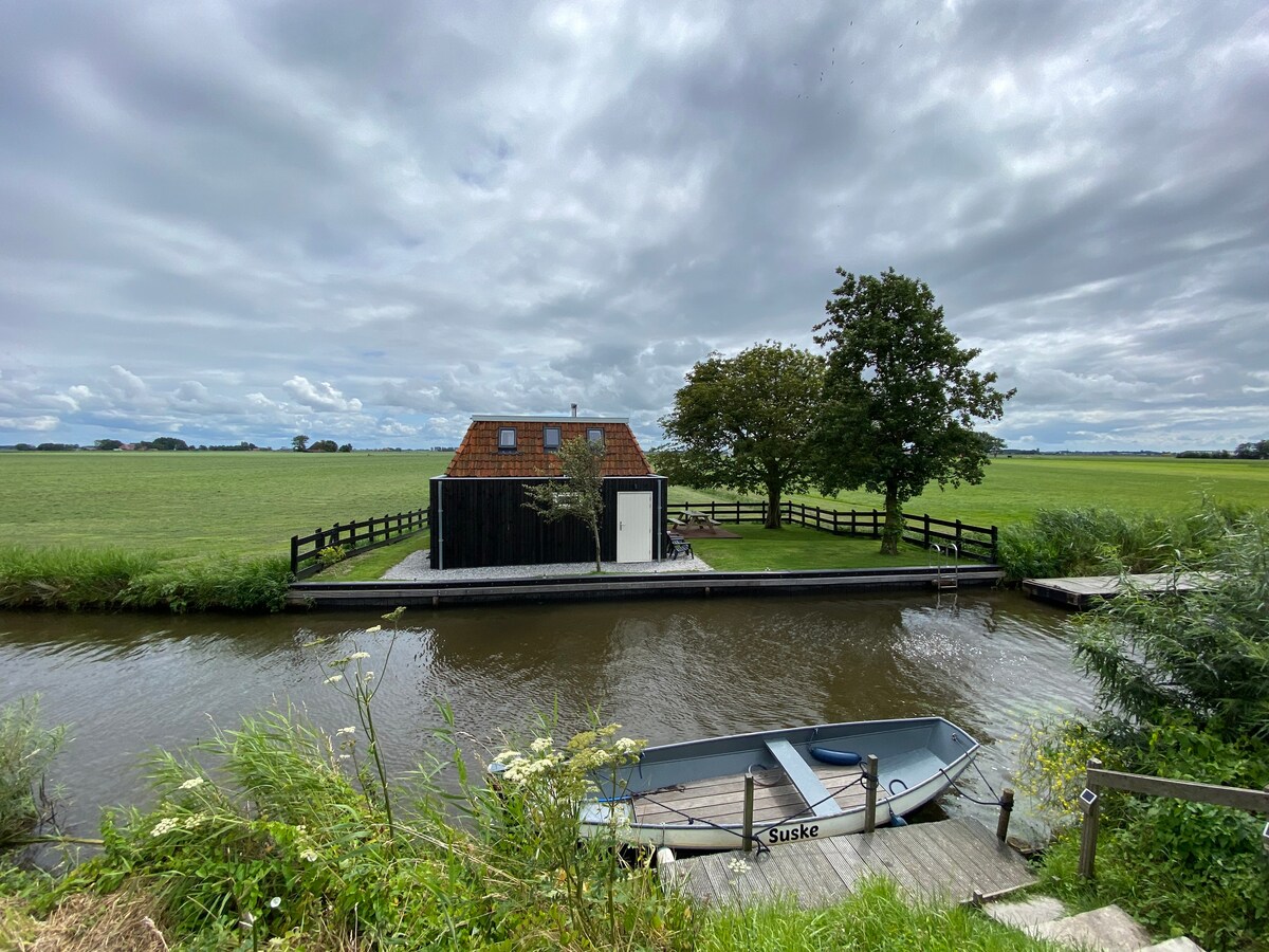 The image captures a serene view of the holiday home standing beside a calm canal. Lush green fields stretch into the distance, while a small boat is moored nearby. A tree offers shade, contributing to the peaceful outdoor setting.