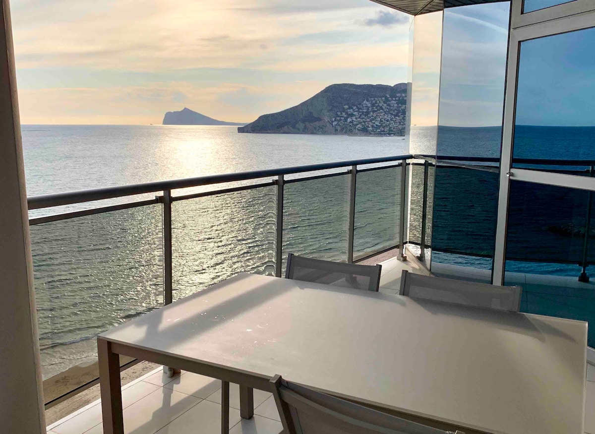 A spacious terrace is presented with a large dining table surrounded by chairs, offering an unobstructed view of the sea and distant coastline. The gentle waves reflect the soft evening light, while the shoreline is visible in the background. The scene conveys a serene atmosphere.