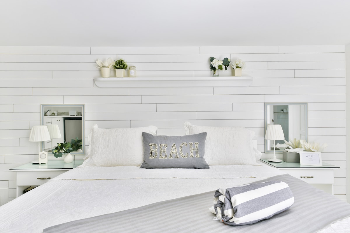 A spacious bed is dressed in white linens, with coordinating pillows and a decorative 'BEACH' cushion at the center. Soft lighting is provided by bedside lamps, complemented by decorative shelves featuring plants. Mirrored accents enhance the inviting and airy atmosphere.