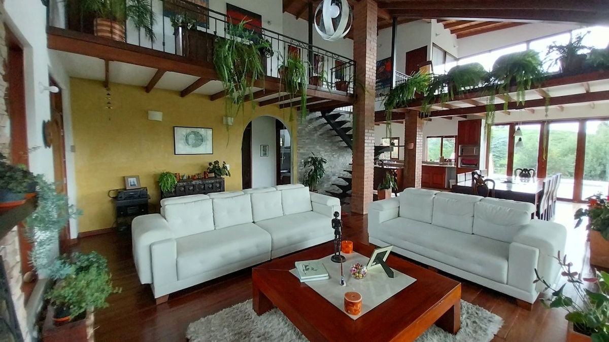 A spacious living area features two white sofas facing a wooden coffee table, surrounded by lush greenery. An intricate staircase leads to the upper level, with large windows allowing natural light to fill the space. Warm wooden accents enhance the room's contemporary design.