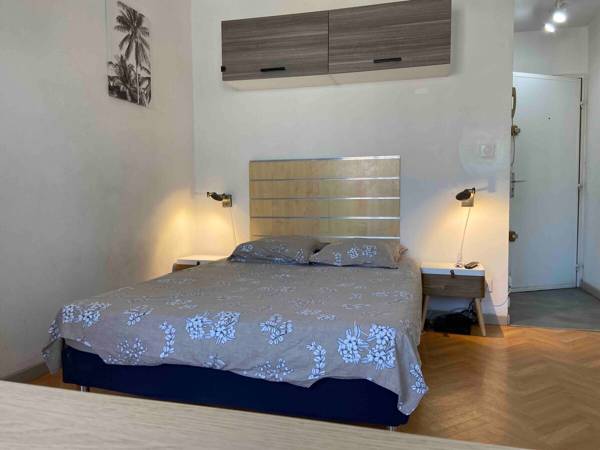 A sleeping area features a spacious bed dressed in a neutral floral-patterned coverlet. Nightstands with lamps are positioned at either side of the bed. A simple artwork with palm trees is displayed on the wall, and the entrance is visible in the background.