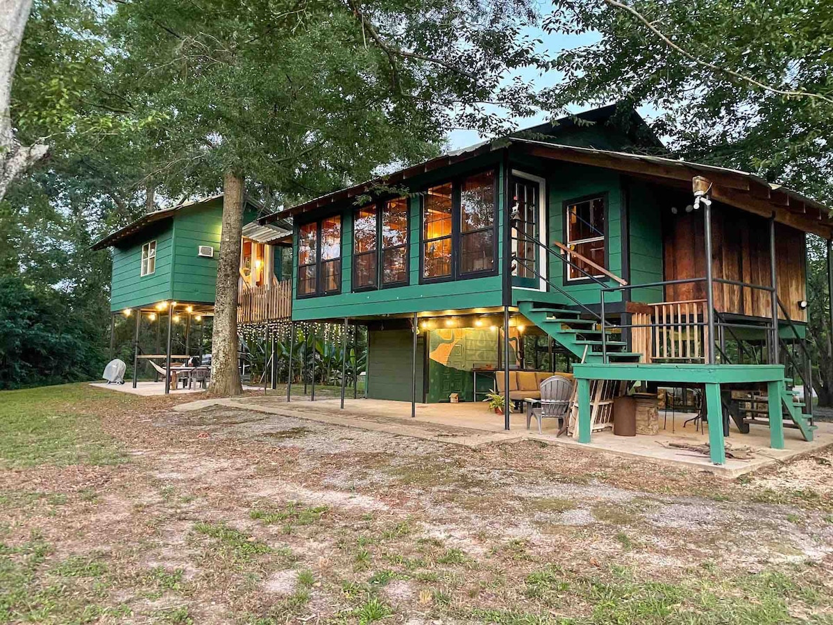 The treehouse features a two-story structure surrounded by greenery. A large deck with multiple windows offers views of the outdoors. Steps lead up to the main entrance, while a cozy sitting area with chairs is positioned nearby. Natural landscaping complements the tranquil setting.