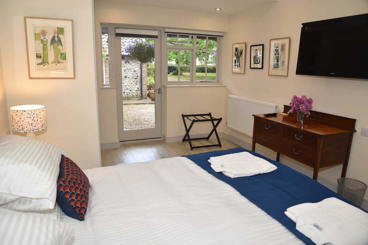 A bright room features a spacious bed with crisp linens and decorative pillows. Natural light floods through large windows, leading to a private outdoor area. Nearby, a wooden dresser displays fresh flowers, while a folding chair offers additional seating.