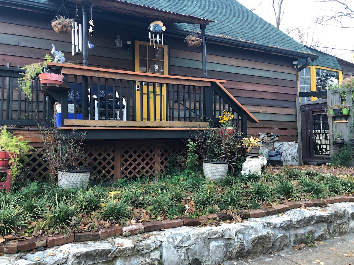 A wooden structure features a welcoming yellow door and a porch adorned with potted plants. Hanging wind chimes add a decorative touch. Lush greenery surrounds the entrance, complemented by stone pathways and a fenced area, creating a quaint and inviting outdoor space.
