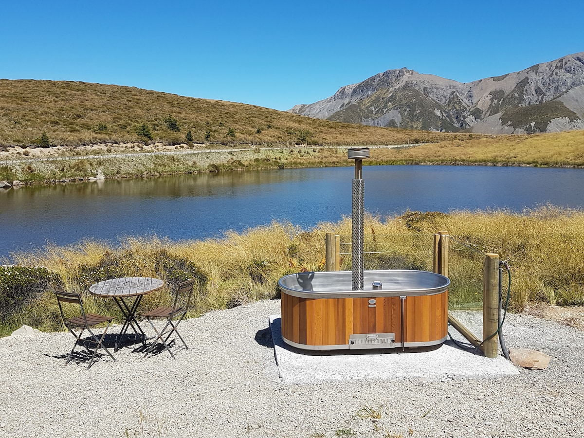 A wood-fired hot tub is positioned beside a tranquil mountain lake, surrounded by golden grasslands and scenic mountain views. A small round table with two chairs sits nearby, creating a serene outdoor relaxation spot under a clear blue sky.