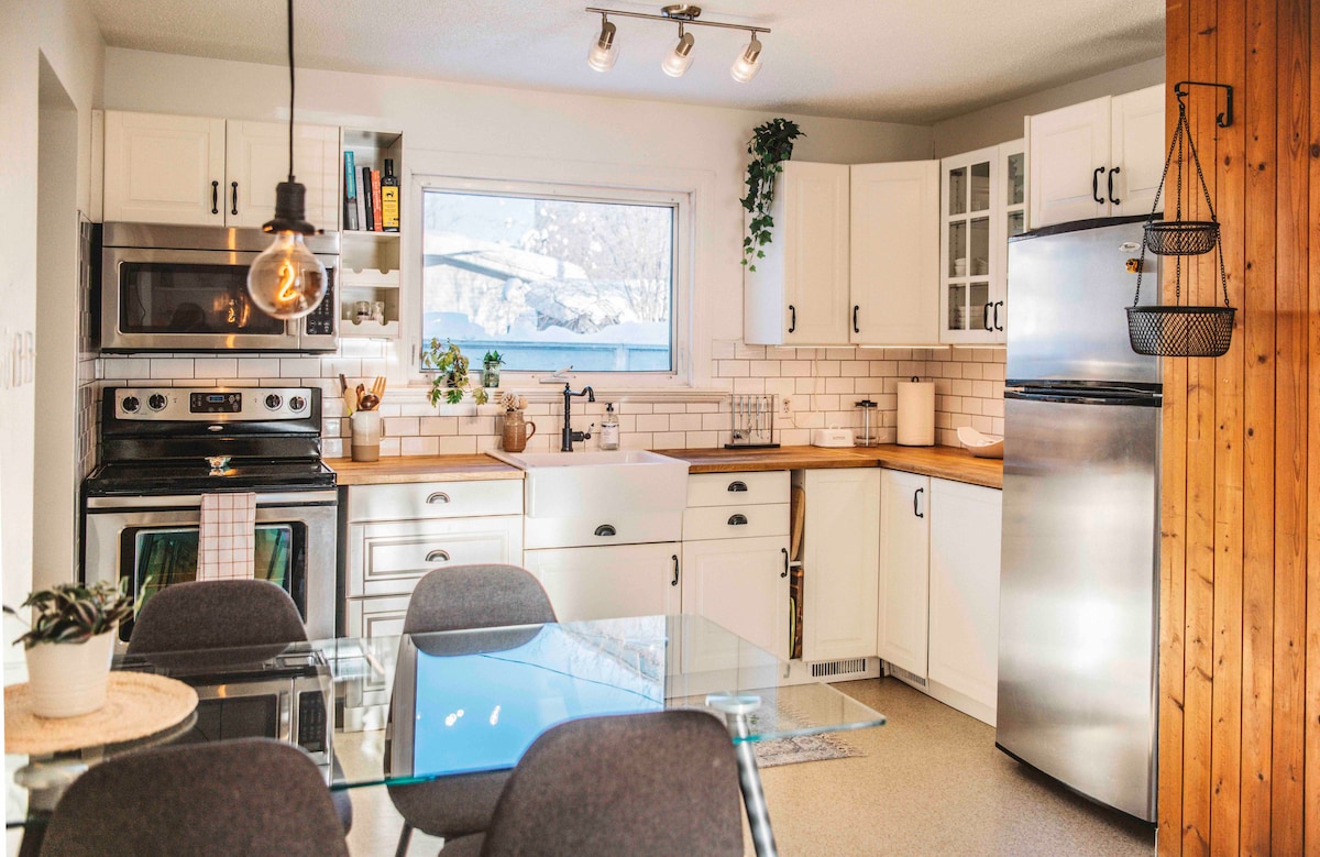 A modern kitchen is showcased with white cabinetry and light wood accents. Stainless steel appliances, including a range and refrigerator, are present. A glass dining table surrounded by four gray chairs sits near a bright window, allowing natural light to illuminate the space.