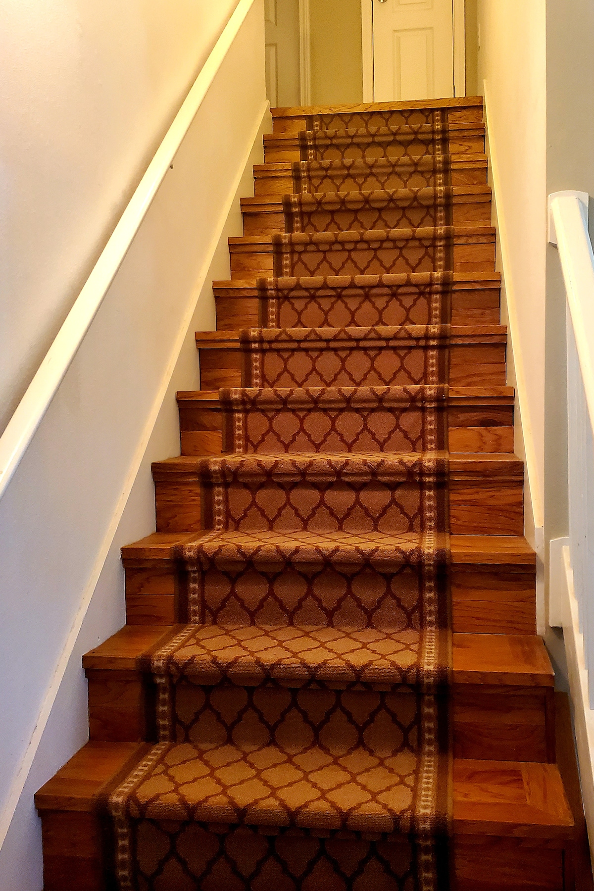 A set of stairs is shown, featuring wooden steps covered with a patterned runner. The handrail is painted white, and light from the upper area illuminates the steps. A door is visible at the top of the stairs, providing access to the suite.