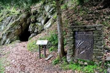 A wooded area features a rocky entrance to a cave, with a stone plaque and informational sign positioned nearby. Surrounding vegetation includes green foliage and fallen leaves. Natural stone elements and soft earth tones enhance the tranquil setting.