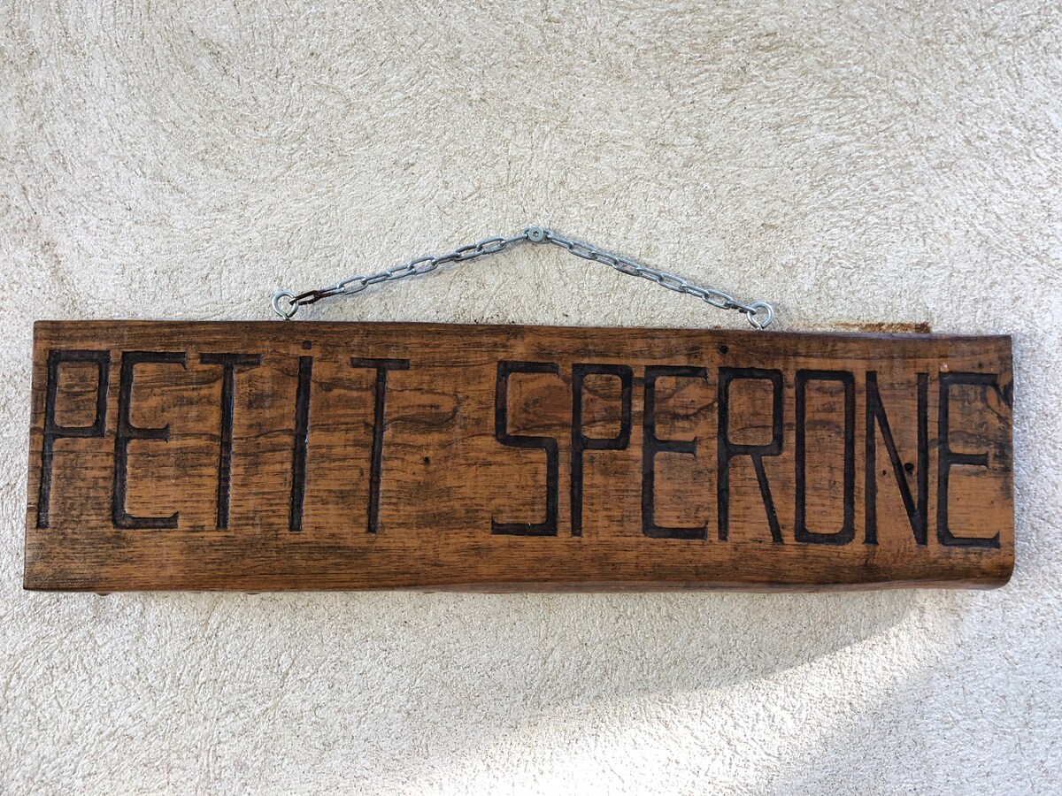 A wooden sign hangs from a chain, displaying the name 'PETIT SPERONE' carved in a bold font. The natural texture of the wood complements the neutral wall background, providing a rustic touch to the property.