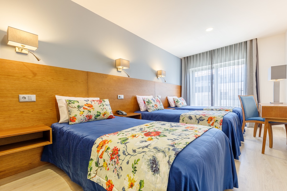 A spacious guest room features three beds with floral patterned bedcovers. Wooden accents provide a warm touch, and soft lighting is offered by wall-mounted lamps. A desk and a chair are positioned to the side, with a window that brings in natural light, dressed in light gray curtains.