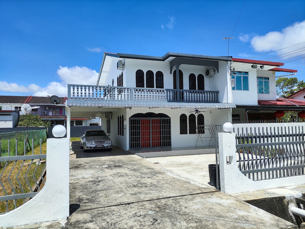 A two-story house is presented with a wide balcony and decorative railings. A concrete driveway is visible, leading to a parked car. The facade features multiple arched windows and a central door, while a gated entrance provides access to the property.
