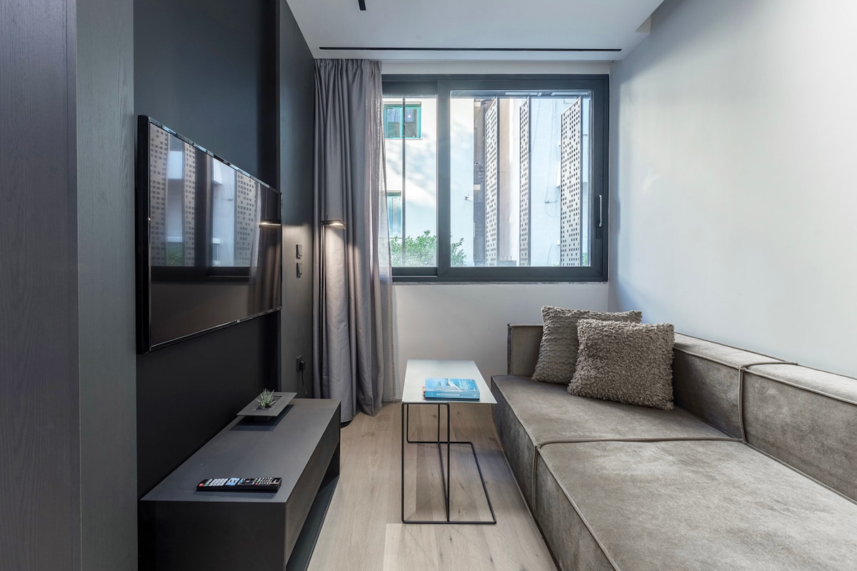A modern living area features a neutral color palette with a plush sectional sofa and a sleek coffee table. A wall-mounted television is positioned opposite a large window, allowing natural light to fill the space while sheer curtains add a touch of privacy.