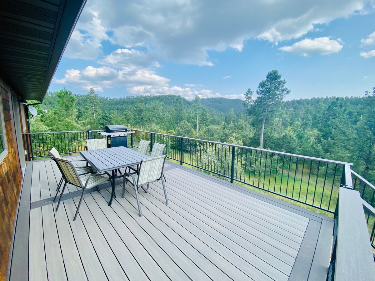 A spacious deck is shown with outdoor seating for six, facing expansive views of the wooded landscape and distant hills. A grill is positioned nearby, complementing the outdoor dining space under a partly cloudy sky.