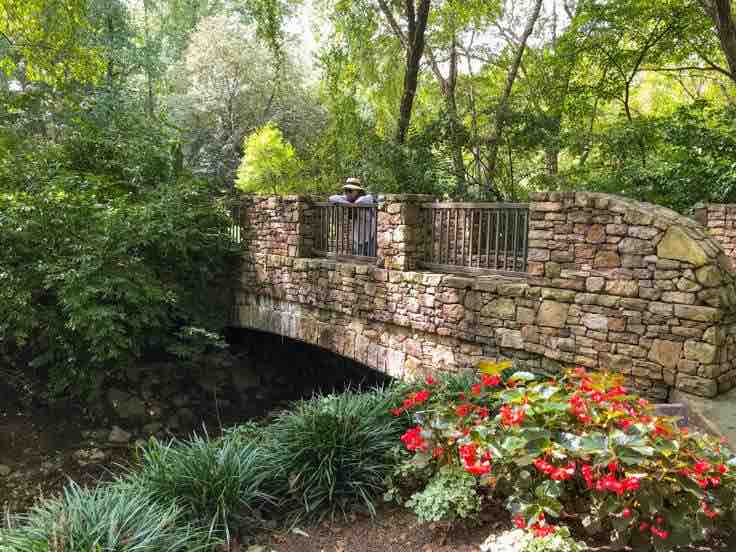 A stone bridge is observed arching over a serene stream, surrounded by lush greenery and colorful blooming flowers. The structure features a detailed stonework design and low protective railings, blending harmoniously into the natural landscape.