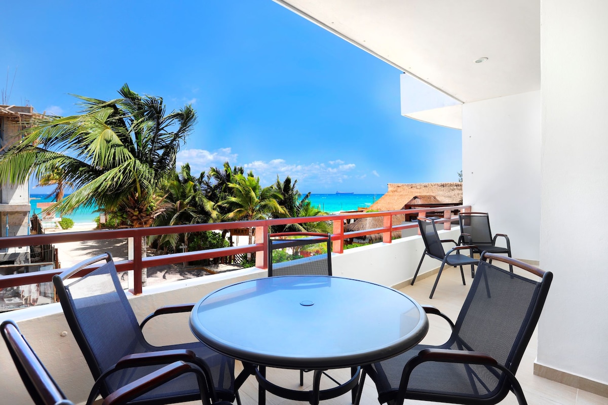 A private terrace is shown, furnished with a round table and four black chairs. The space offers a partial view of the shimmering Caribbean Sea and lush palm trees, inviting relaxation and outdoor enjoyment.