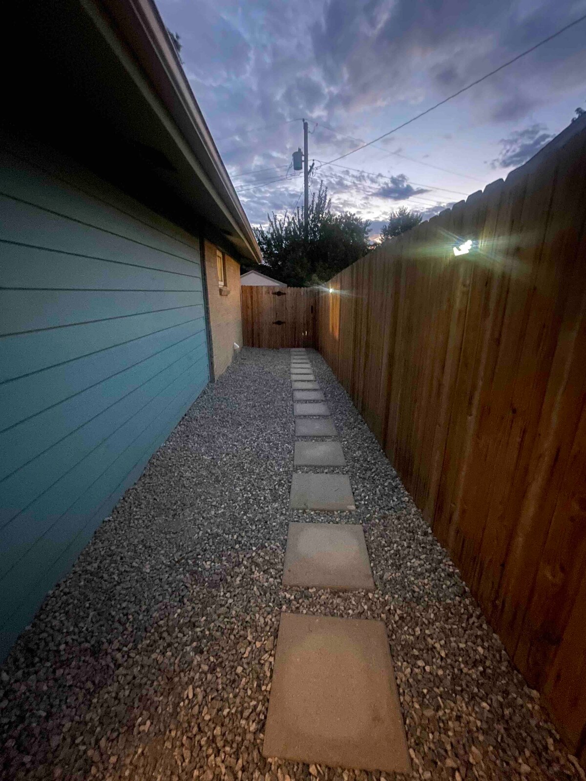 A pathway lined with stepping stones leads alongside the side of the house. The area is bordered by a wooden fence and features gravel landscaping. Soft lighting illuminates the path, creating a welcoming ambiance as dusk settles in.