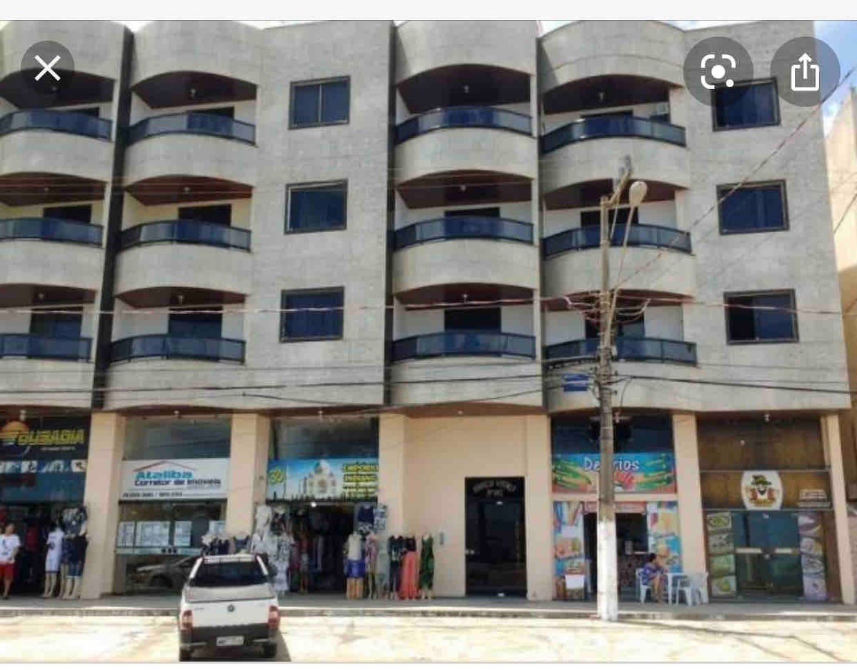 A modern building features multiple balconies with dark railings, extending across the upper levels. The ground floor displays several storefronts with vibrant signage, while a white vehicle is parked in front of the establishment. Street utilities are visible, establishing a bustling urban environment.