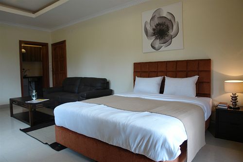 A cozy bedroom features a spacious bed with crisp white linens and a neutral-colored blanket. A large flower artwork hangs above the bed. A comfortable sofa sits beside a dark wooden coffee table, with a lamp providing soft illumination.
