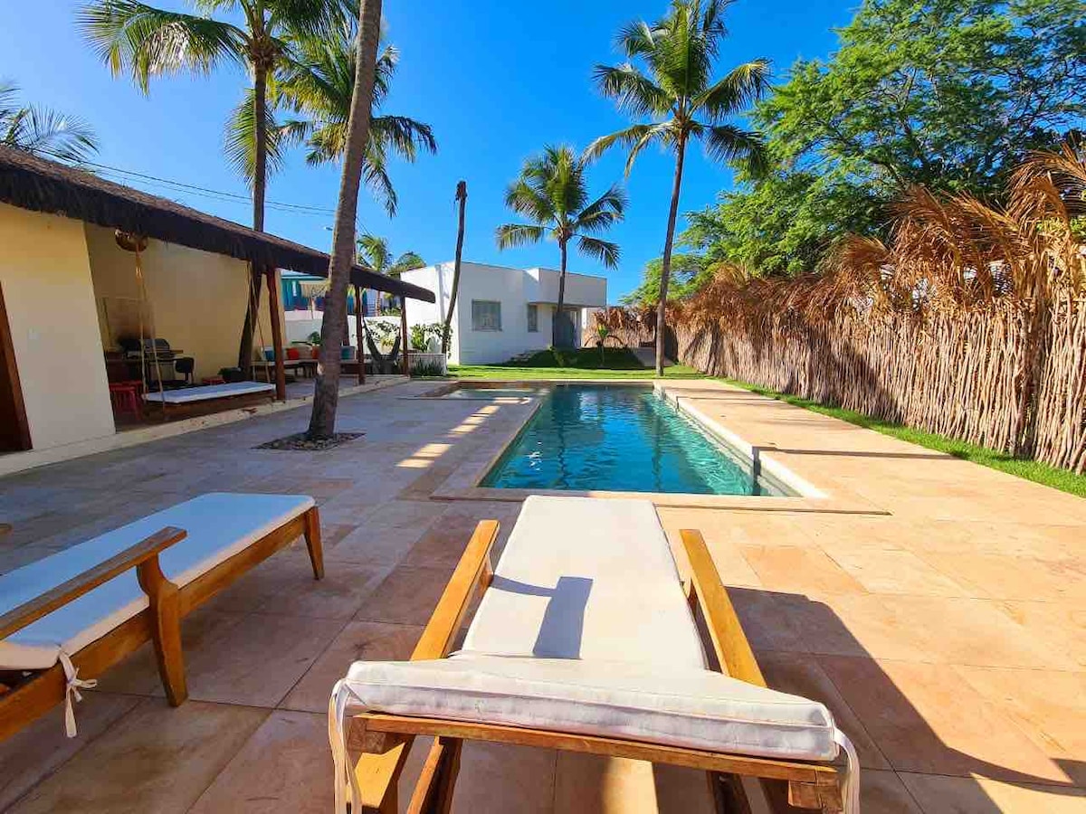 The outdoor space features a private swimming pool surrounded by a spacious tiled patio. Sun loungers are placed nearby, with palm trees and natural vegetation offering a tropical feel. The clear blue sky provides bright sunlight, creating an inviting environment.