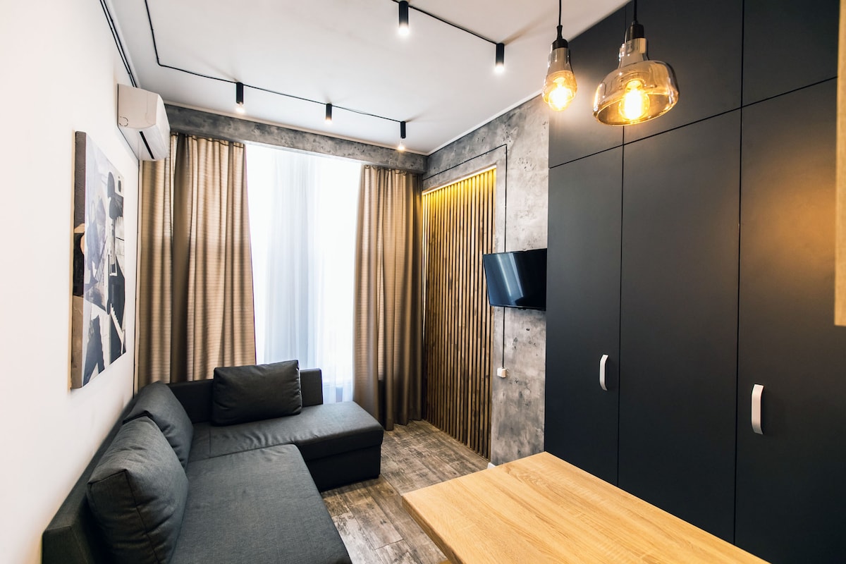A modern living space features a gray sectional sofa positioned beside a small wooden table. Natural light streams through floor-to-ceiling curtains, illuminating the wall with decorative panels. A flat-screen television is mounted on the dark cabinet, complementing the overall sleek design of the room.