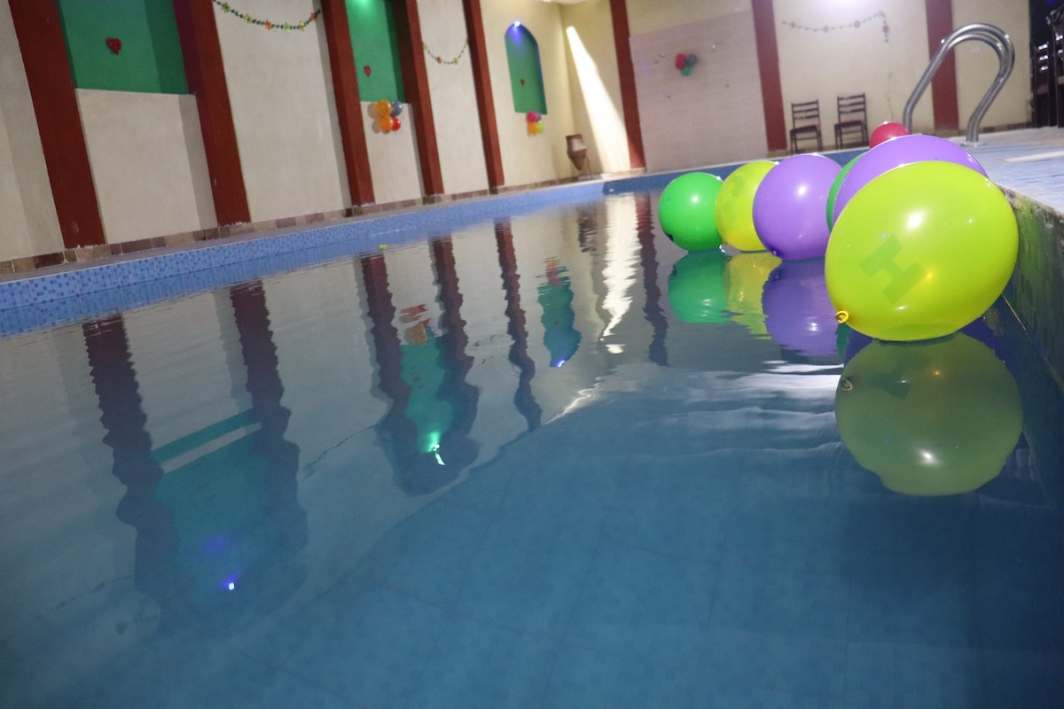 The image features a swimming pool with a clear blue surface reflecting colorful balloons floating on the water. The pool area is enhanced by soft lighting and bright walls, creating a cheerful environment suitable for relaxation or gatherings.