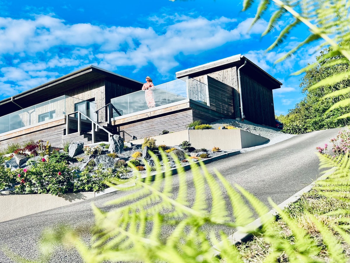 A modern, wooden cottage is situated on a hill, featuring a glass railing on the balcony. The driveway slopes gently upwards, surrounded by vibrant flowers and lush greenery. A person stands on the balcony, enjoying the view under a partly cloudy blue sky.