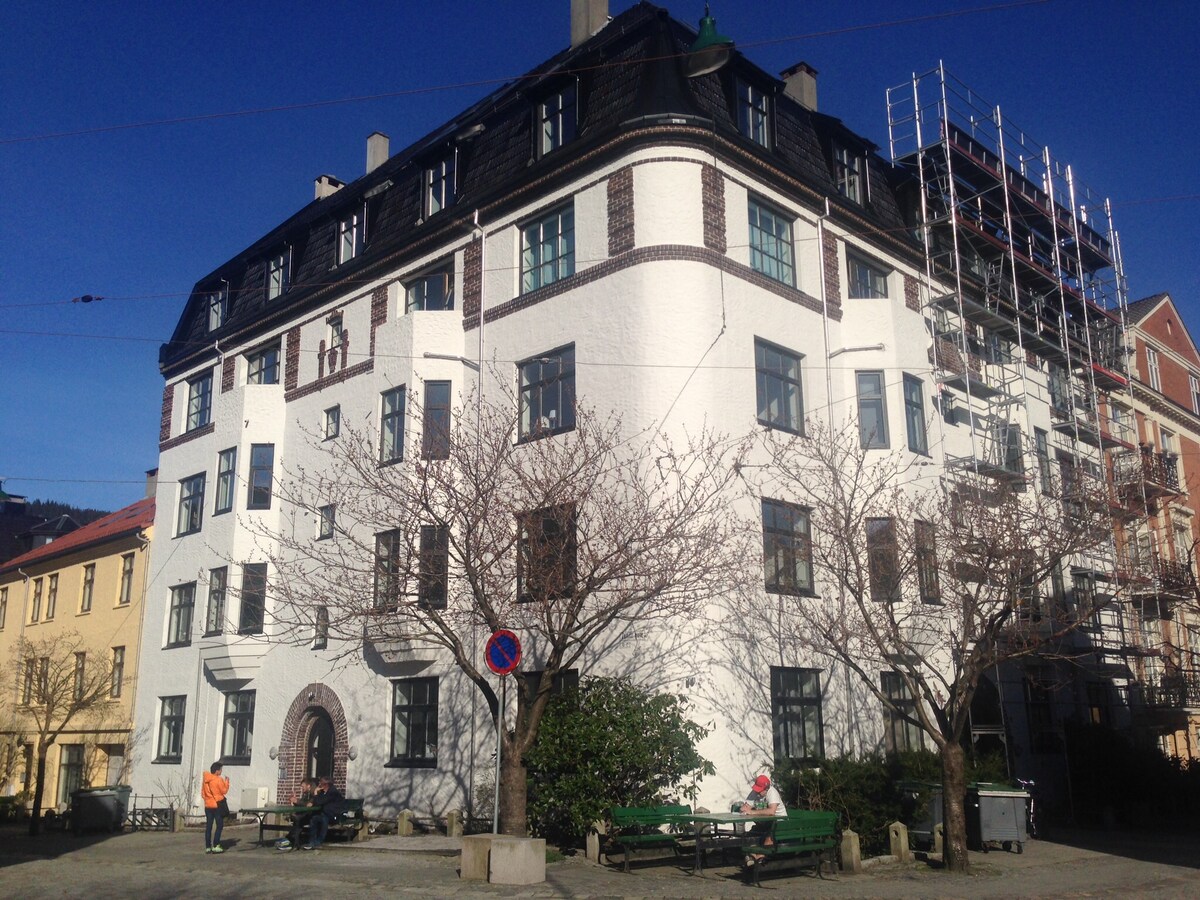 An elegant building features a white facade with intricate detailing and a distinctive roofline. Surrounding trees provide shade, while benches are positioned nearby for relaxation. Construction scaffolding can be seen on one side, indicating ongoing work. A clear blue sky enhances the overall scene.