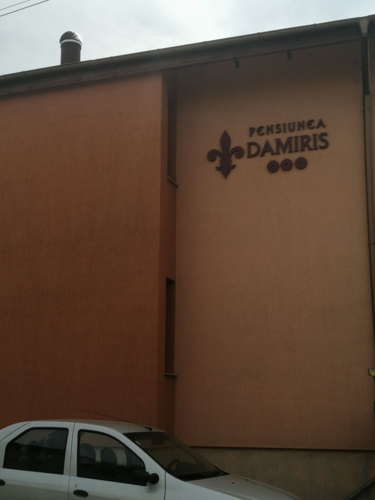 The exterior of Pensiunea Damiris is captured, displaying a soft-colored building facade featuring the name of the guesthouse prominently above a modestly parked vehicle. The structure exhibits clean lines and a simple architectural design, contributing to its welcoming presence.