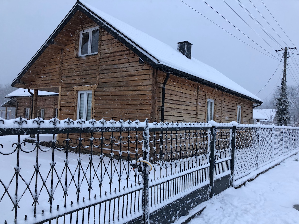 A wooden house with a sloped roof is shown, surrounded by a snow-covered landscape. The structure features large windows that allow natural light in. A decorative metal fence lines the front, adding to the rustic charm of the setting.
