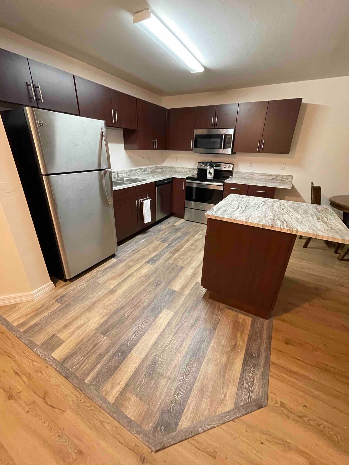 The kitchen features modern cabinetry with dark wood finishes and a light granite countertop. A stainless steel refrigerator and stove are included, along with a microwave. The spacious layout allows for easy movement, complemented by a wood-style floor that adds warmth to the room.