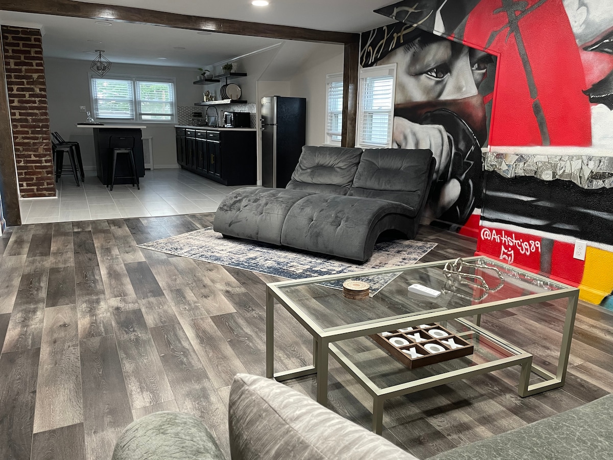 A spacious living room features a sleek grey sectional sofa positioned near a decorative area rug. A contemporary coffee table with a decorative centerpiece accompanies the seating area. A vibrant mural adorns the wall, complemented by a well-organized kitchenette visible in the background.