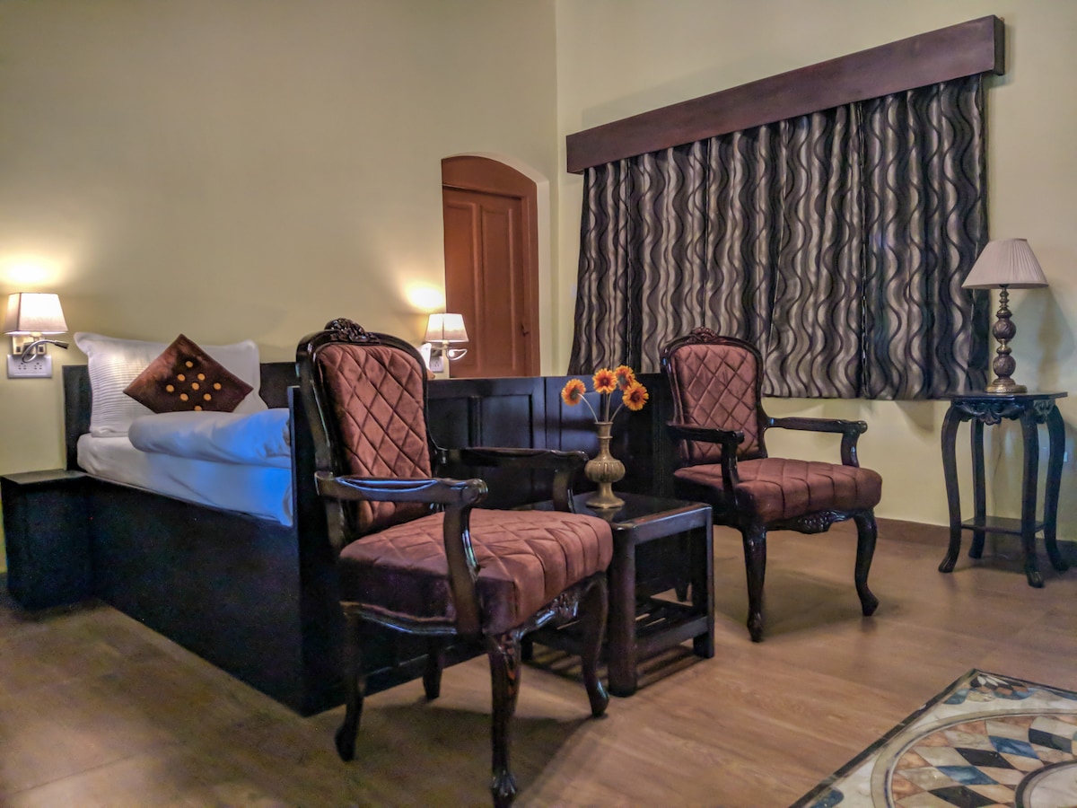 The room features an elegant teak bed and two intricately carved chairs positioned around a small table. A vase of sunflowers adds a touch of color. Natural light is filtered by patterned curtains, creating a cozy atmosphere.