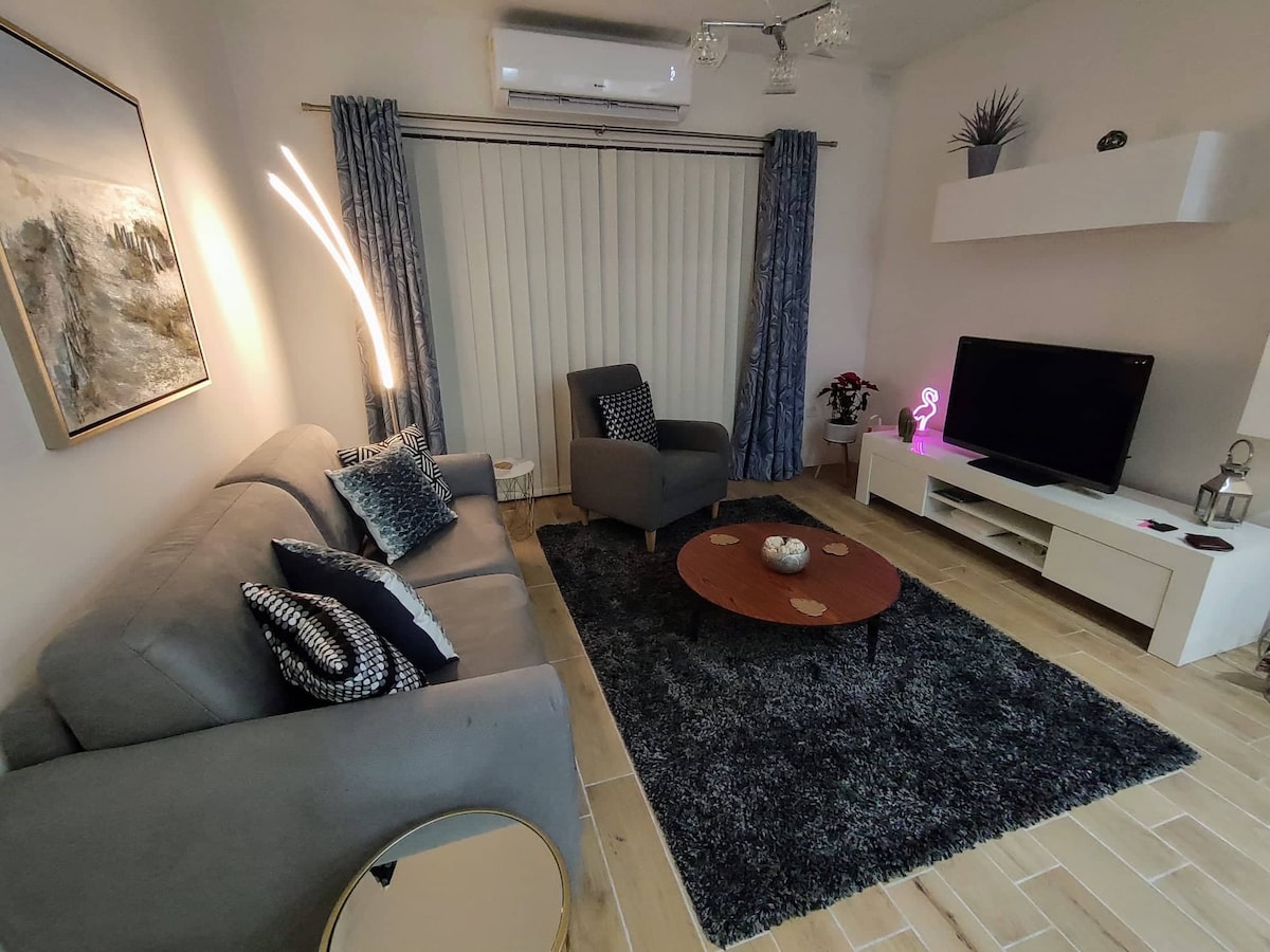 A cozy living area features a plush grey sofa adorned with decorative cushions. A round wooden coffee table sits at its center, surrounded by a soft area rug. A modern armchair occupies one corner, and a wall-mounted TV stands opposite, framed by elegant curtains.