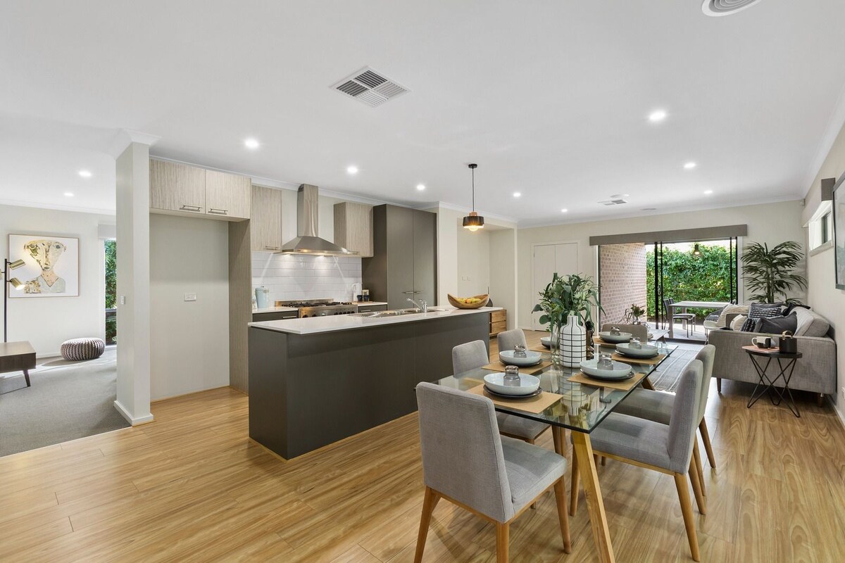 A modern open-plan living space is captured, featuring a sleek kitchen with stainless steel appliances and a dining area with a glass table set for meals. Neutral tones dominate the decor, complemented by natural light from large windows leading to a lush outdoor area.