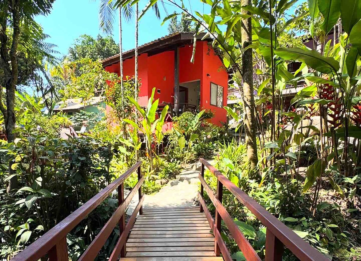 A wooden bridge leads through lush greenery to a vibrant orange chalet nestled among tropical plants. Sunlight filters through the leaves, illuminating the pathway and enhancing the natural setting surrounding the accommodation.