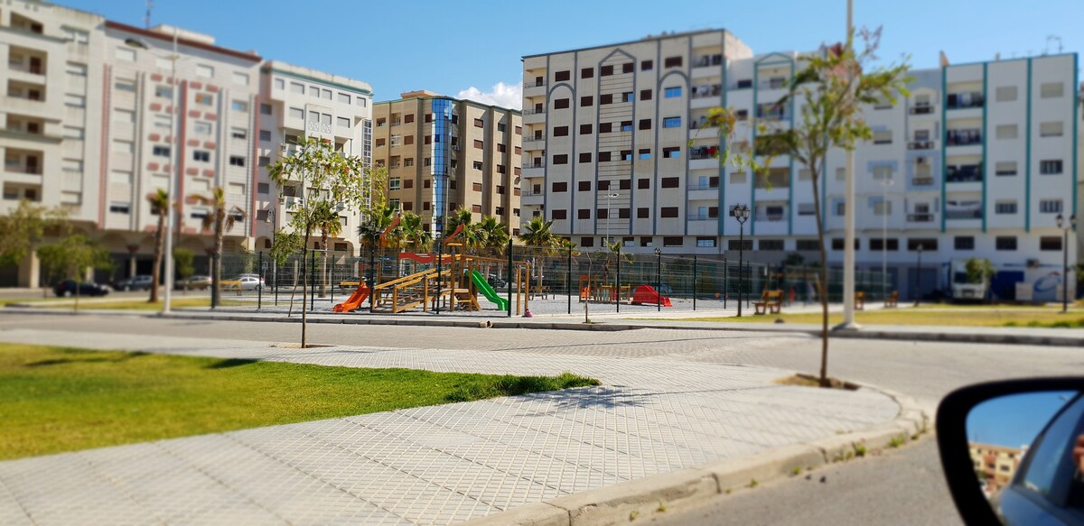 A neighborhood playground is visible, featuring bright orange and green play structures surrounded by well-maintained grass. Surrounding residential buildings reflect a modern design with multiple stories. The sky is clear, allowing natural light to illuminate the open space.