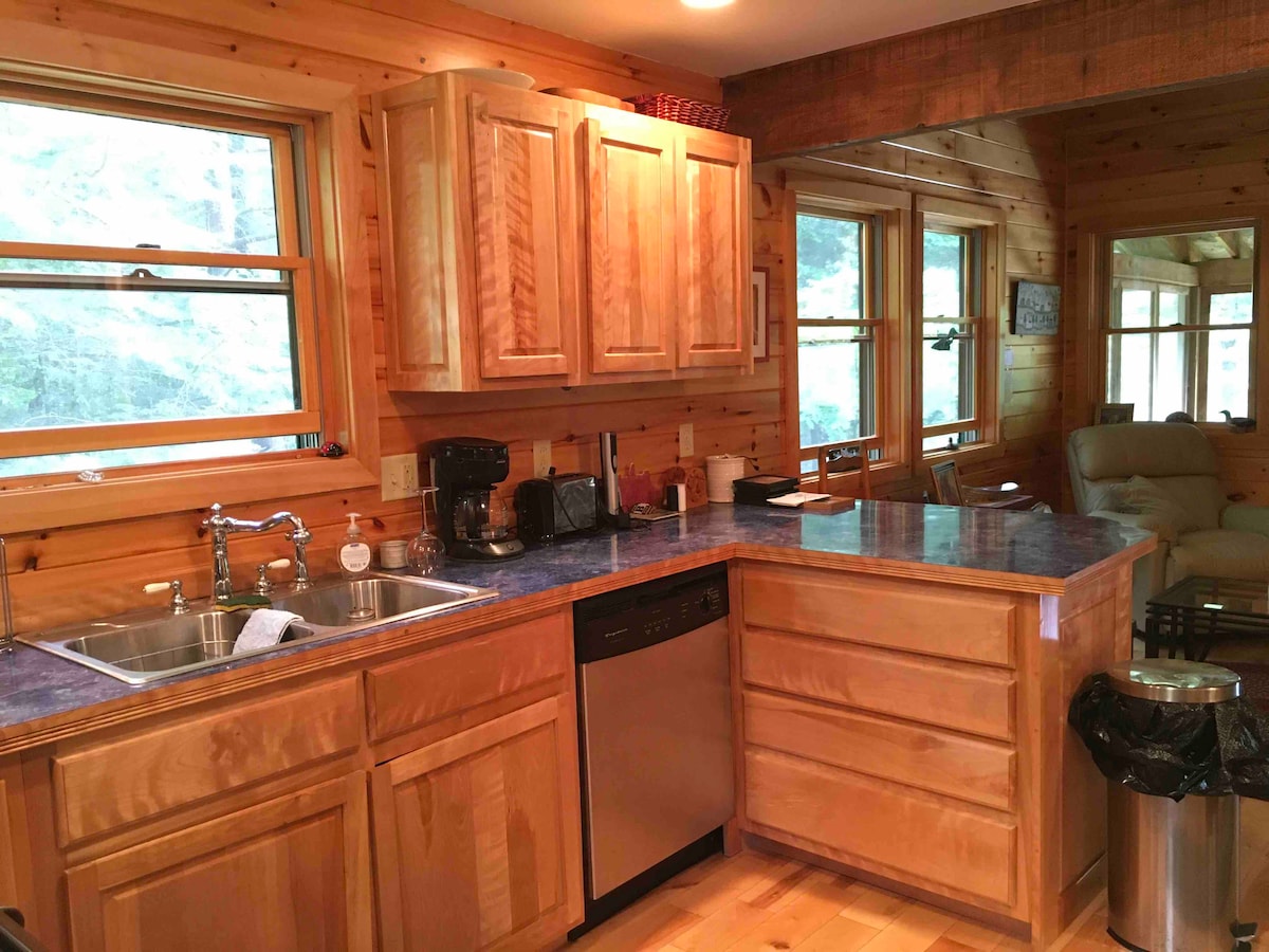 A wooden kitchen area features warm cabinetry and a blue countertop. Modern appliances, including a dishwasher and coffee maker, are present. Sunlight filters through multiple windows, illuminating the space and providing views of the surrounding nature.