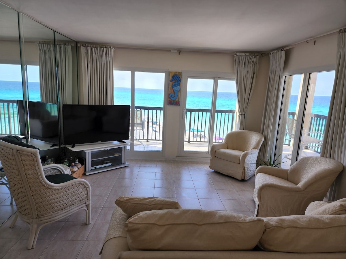 A bright living area features large glass doors that open to a balcony with expansive beach views. Two light-colored armchairs and a wicker chair complement a modern television cabinet. The walls are adorned with mirrors, reflecting natural light and the serene coastal surroundings.