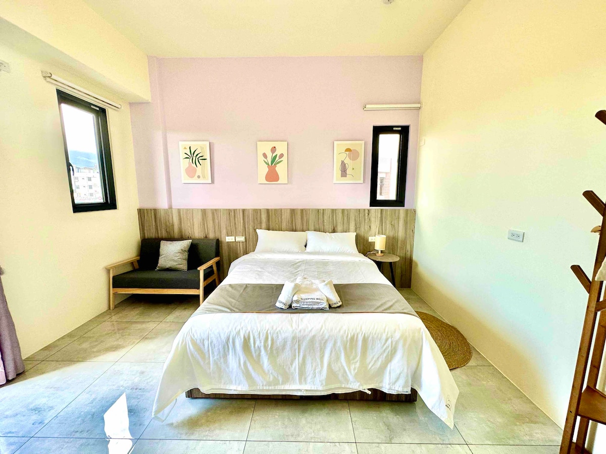 A spacious bedroom is furnished with a large bed dressed in white linens and soft gray accents. Adjacent is a cozy seating area with a dark couch. Light enters through two windows, enhancing the calming atmosphere created by pastel wall art.