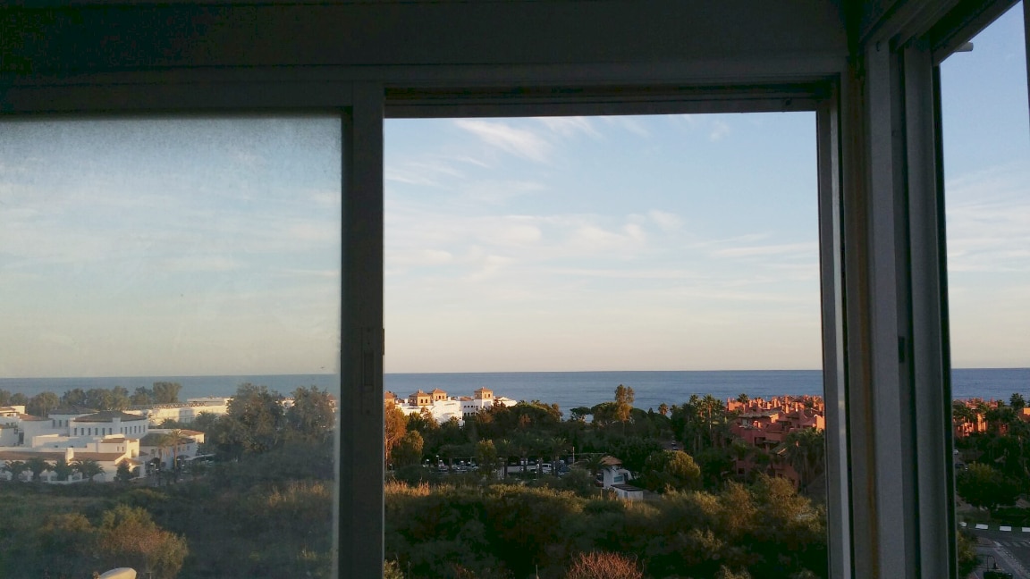A view from a window presents the sea and surrounding landscape. The horizon blends with the sky, while buildings and greenery are seen below. The glass window allows natural light to fill the space, emphasizing the scenic coastal environment.