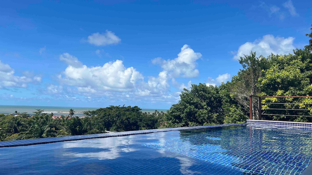 The infinity pool creates a seamless view of the horizon, where the ocean meets the sky. Lush greenery surrounds the area, enhancing the sense of tranquility. The clear blue water reflects the clouds and sunlight, inviting relaxation and calm.