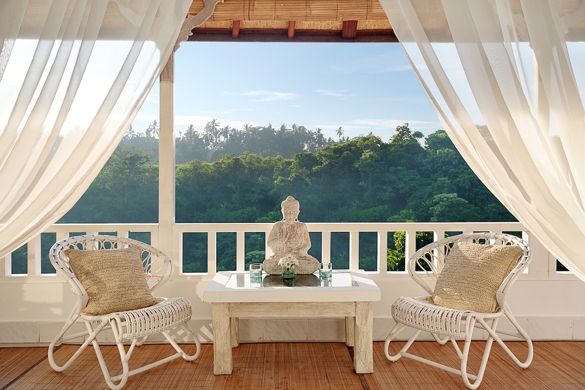 A private balcony features two woven chairs and a white table, creating a serene space. A statue is placed centrally among a decorative arrangement. Sheer white curtains gently frame the view overlooking the lush jungle canopy, inviting natural light into the area.