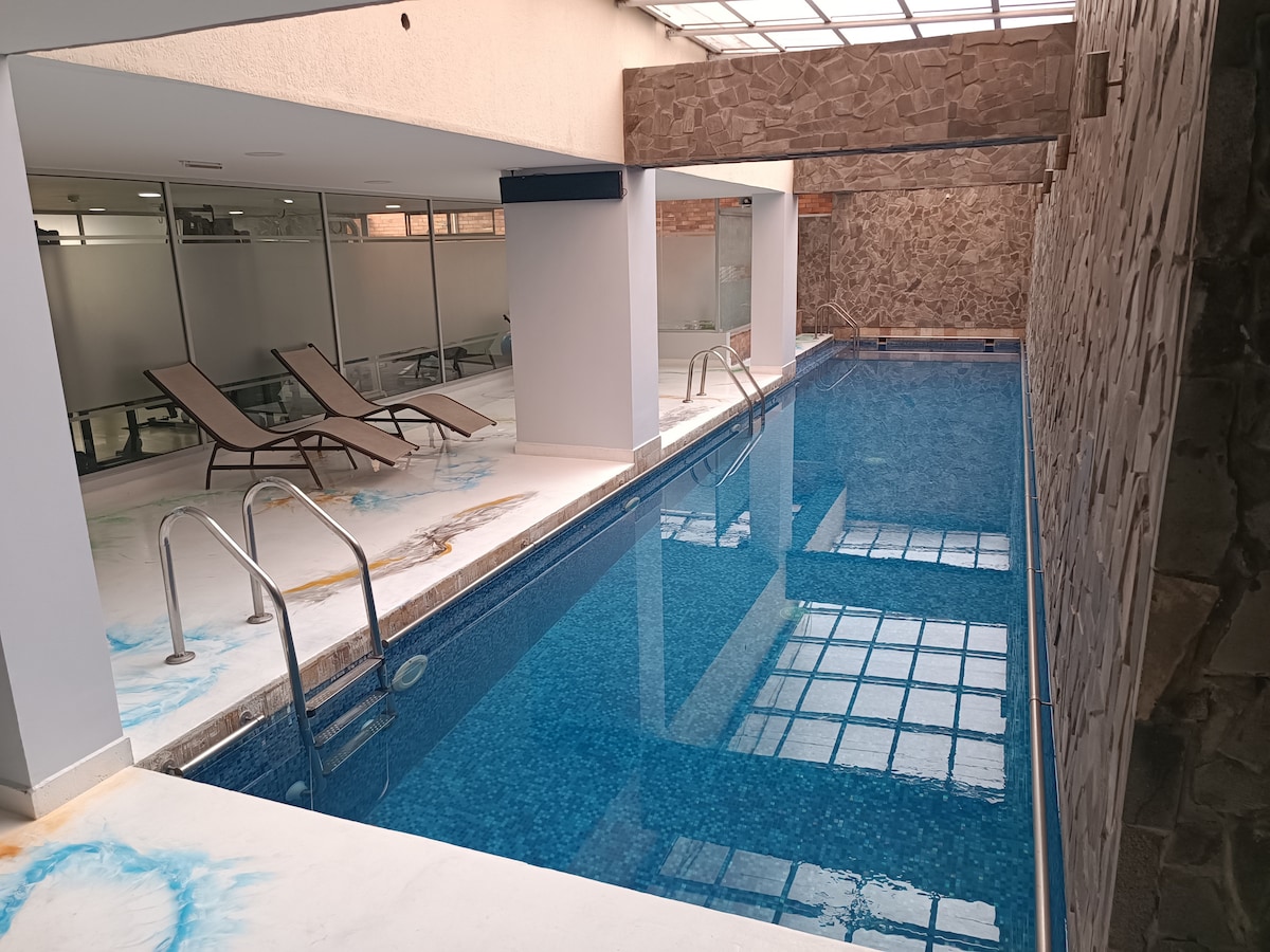 An indoor swimming pool is depicted, featuring clear blue water and a tiled surface. Loungers are placed beside the pool, and large windows allow natural light to fill the space. The walls are finished with a mix of smooth and textured materials.